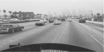 Photograph shows a road view from a car. Many cars are shown on the road and the buildings on the background appear dim due to the smog.
