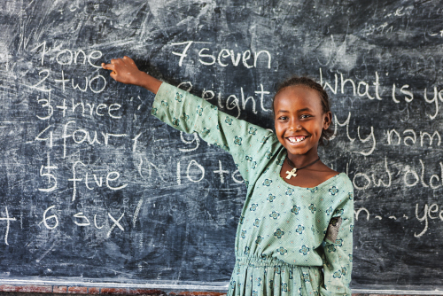 Photo showing young girl child standing near the blackboard. Her right hand is pointing to the word “one” on the board.