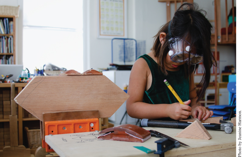 Photo displaying a young girl wearing protective goggles while holding down a piece of wood and using a pencil.