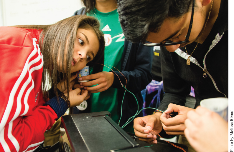 Photo of 3 students in Tanya Kryukova’s physics class at Lighthouse Community Charter School, in Oakland, California; man with eyeglasses holding electrical wires of speaker and woman holding a part of speaker near her right ear.