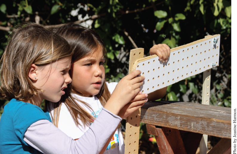 Photo displaying 2 young girls holding up a wooden board that has been drilled with a series of holes and aligning it to the holes on a similarly drilled wooden plank.