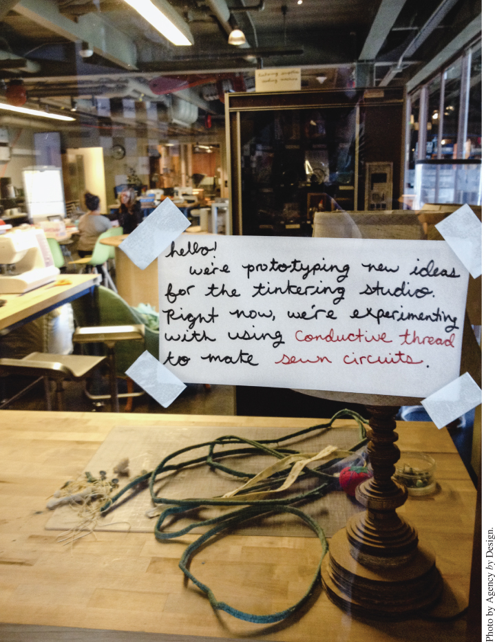 Photo displaying a note posted in a glass window with 2 persons sitting inside the room full of various tools and equipment.