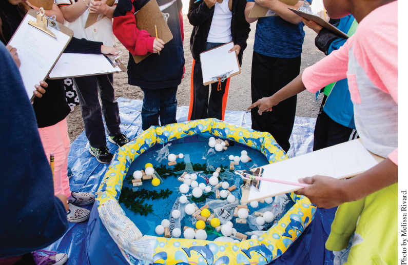 Photo displaying students holding clipboards gathered around an inflatable pool filled with an assortment of materials. One of the students is gesturing at the pool.