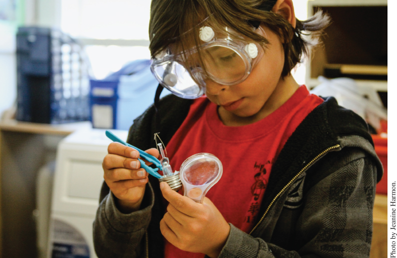 Photo displaying a young boy wearing protective goggles manipulating the internal parts of a light bulb with a tool on one hand while holding the bulb and a magnifying glass on the other hand.