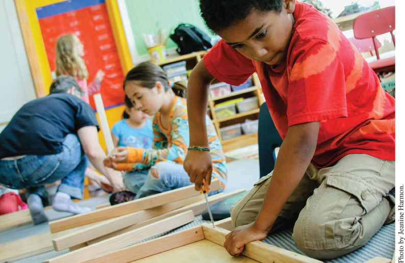 Photo displaying students seated on the floor while handling pieces of wood. A student uses a screwdriver to fasten a screw onto a wooden frame in the foreground.