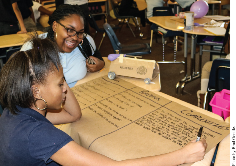 Photo displaying a girl writing on manila paper while another girl, smiling, observes.