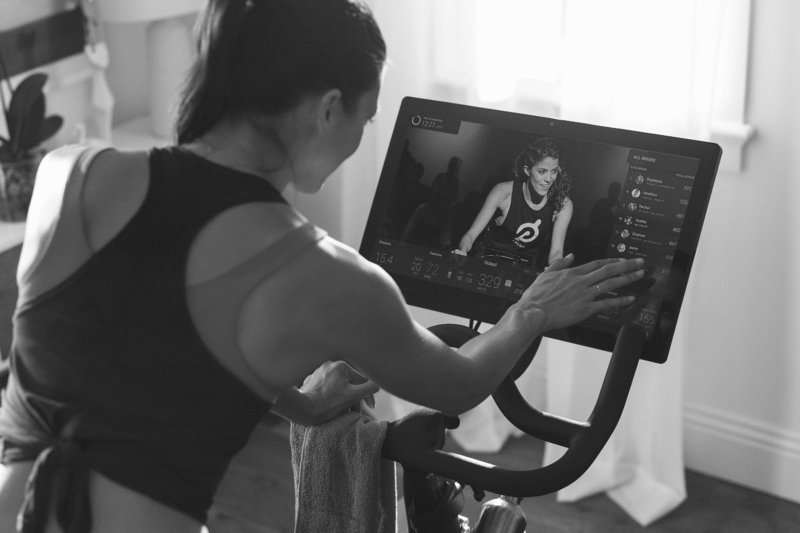 An illustration shows home exercise equipment whose brand is Peloton with a girl working out on the equipment. 