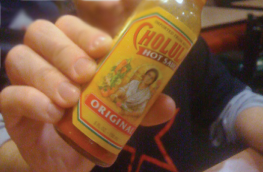 Photograph of the right hand of a person holding a bottle of original hot sauce of a particular brand.