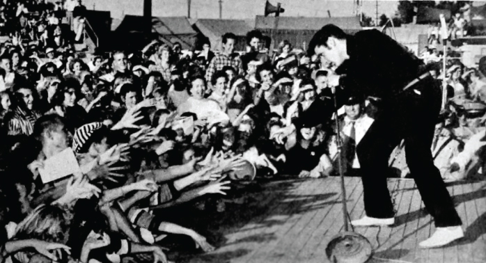 Photograph of a man holding a long mike, singing and reaching out to the audience who want to shake hands with him.