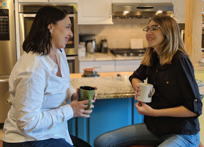 Photograph of an older woman and a girl seated opposite each other, holding coffee mugs, chatting and smiling happily.