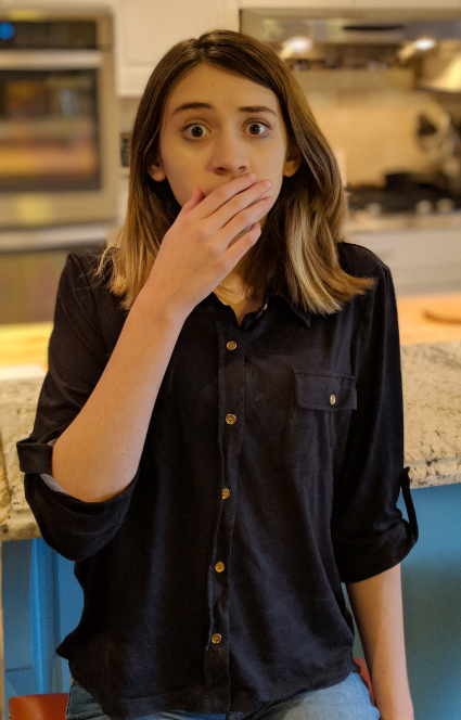 Photograph of a girl standing still with a shocked look on her face, covering her mouth with her right hand.