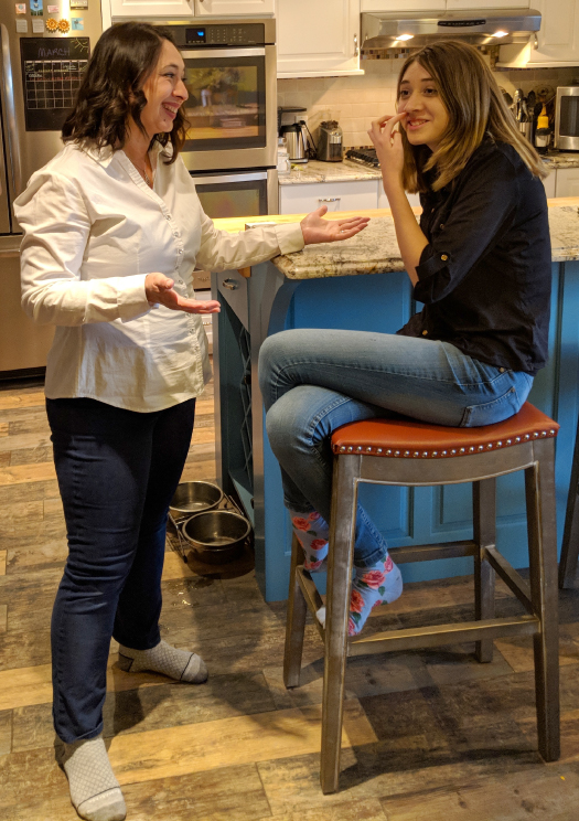 Photograph of a woman standing and happily chatting with a girl seated on a high stool with crossed legs.