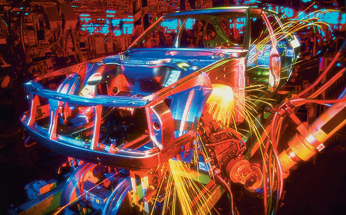 Photograph shows an automobile frame is welded placed on a robotic automatic assembly line.