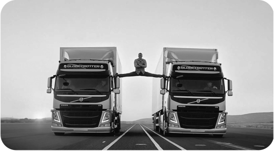 Figure depicting an ad for Volvo where a man with crossed arms is precariously perched on the side mirrors of two moving Volvo trucks, one foot on one truck and one on the other.