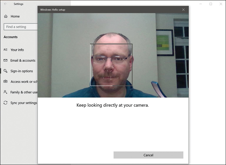 This screenshot shows the Windows Hello setup window with the Settings App in the background. In the center of the window is a picture of a face with several dots or markers overlaid on the image. Below the image, the text Keep Looking Directly At Your Camera appears. Below this text is a Cancel button.