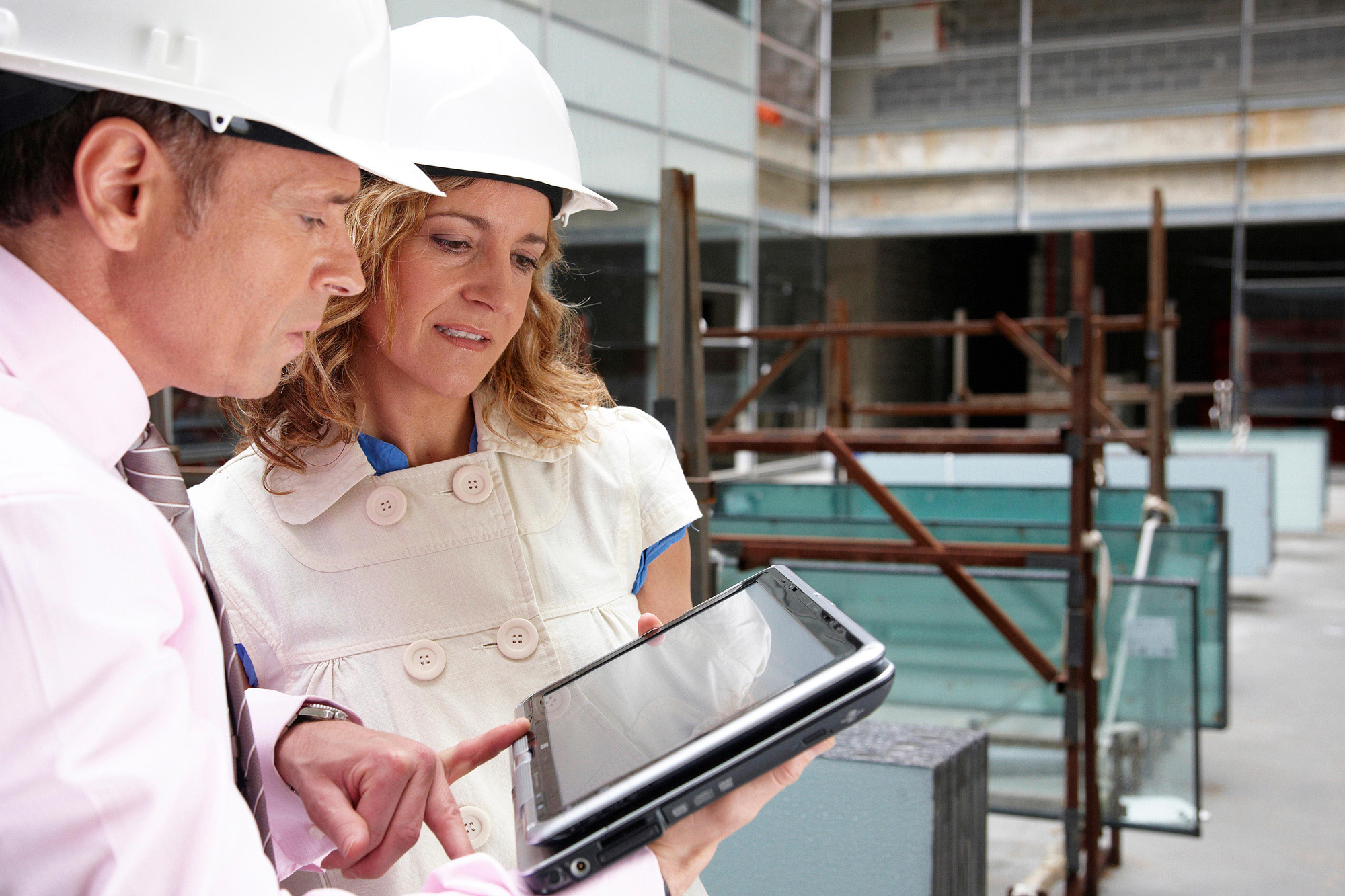 A photo shows a man and woman looking at a tablet computer. They are at a construction site and wear a hard hat.