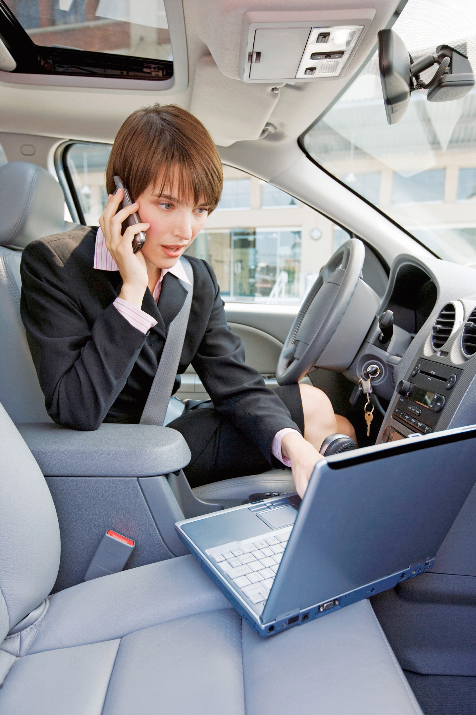 A photo shows a formally dressed young woman speaking over the phone while accessing a laptop inside a car.