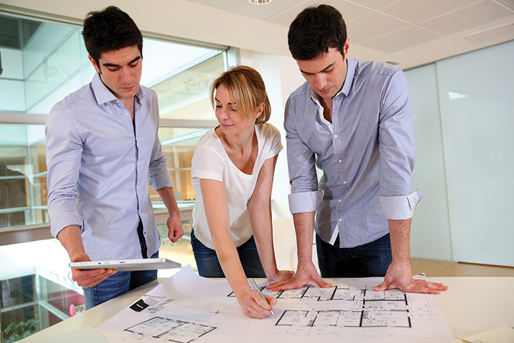 A photo shows a woman and two men studying a blueprint laid on the table, while also checking from a tablet computer.