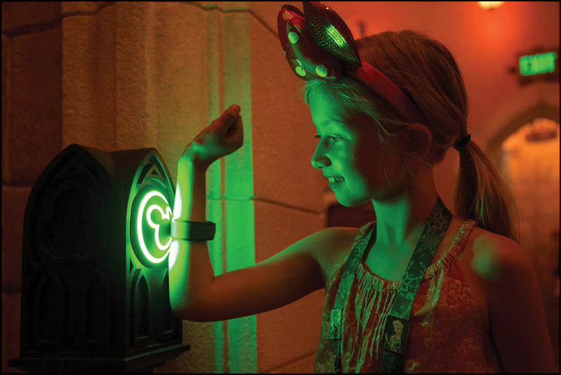 A photo shows a young girl showing her MagicBand RFID wristband to a detector.