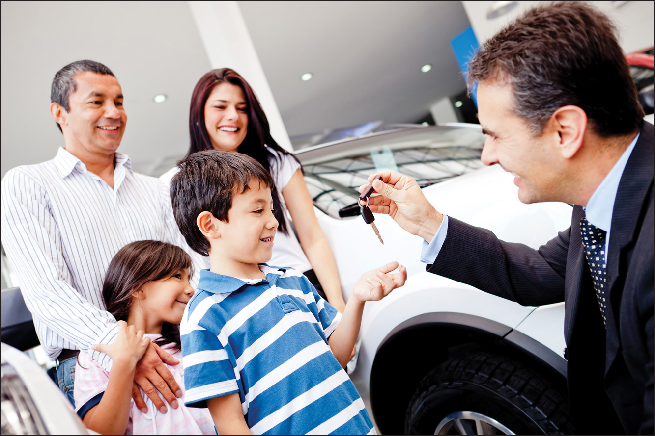 A photo shows a well-dressed man handing over the keys of a car to a young boy in an automobile showroom, while the boy's young sister and his parents look on.