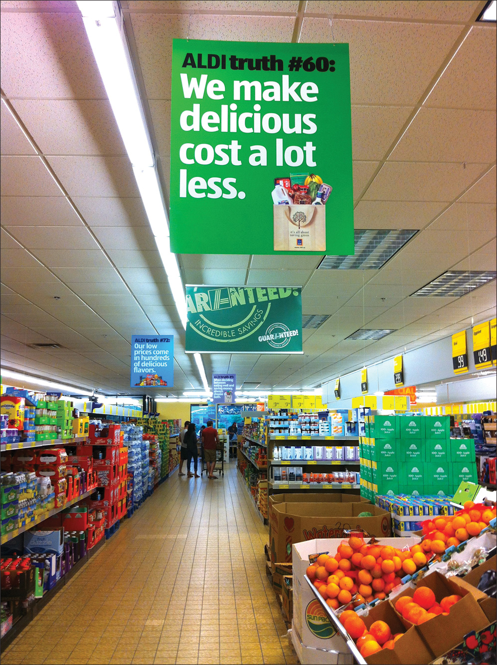 Photo shows an aisle of a supermarket. A banner hanging from the ceiling reads “ALDI truth number 60: We make delicious cost a lot less” along with the photo of a shopping bag that is overflowing.