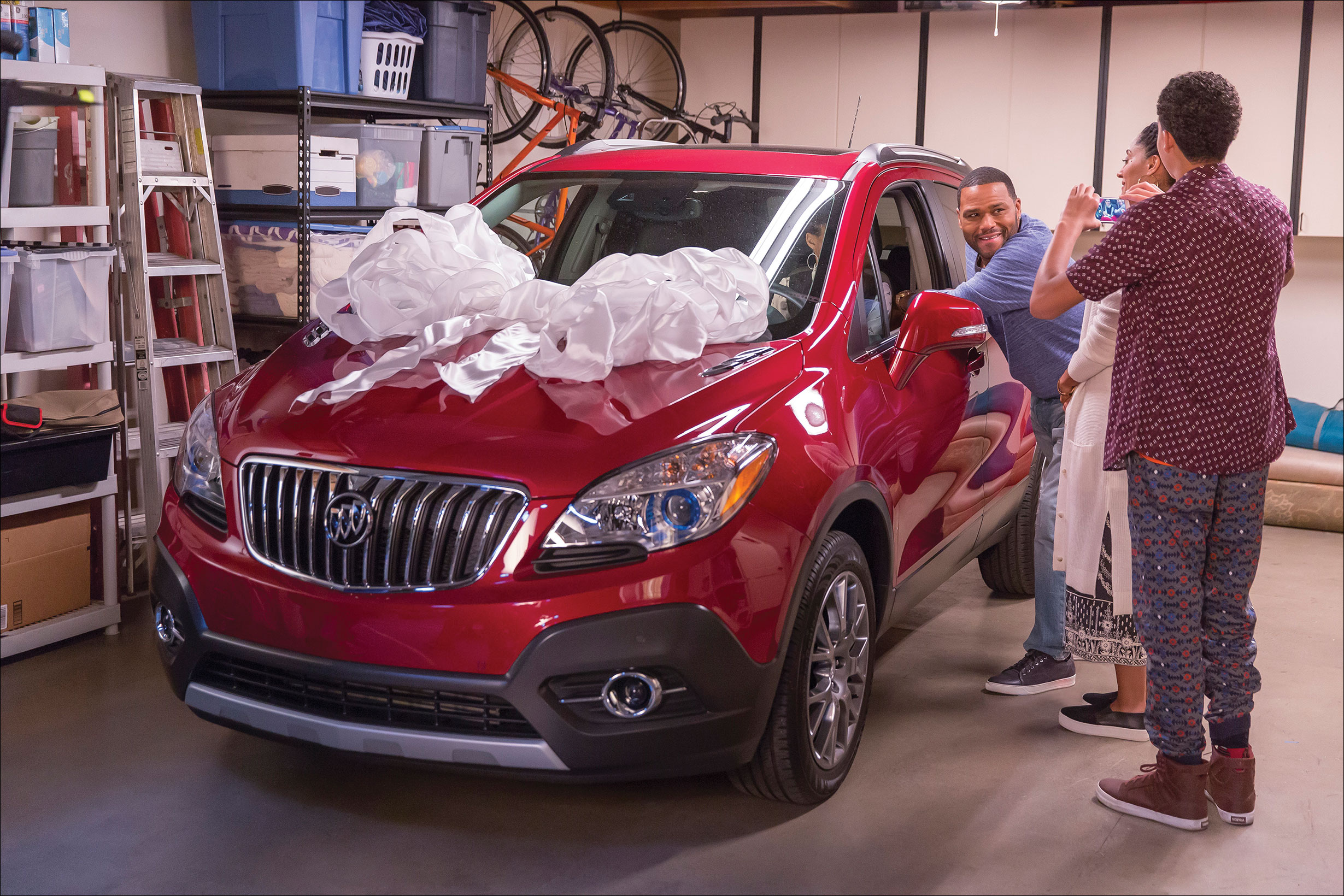 Photo shows a man leaning against the window of a car in a garage, while a man takes his photo on his mobile while a woman looks on.