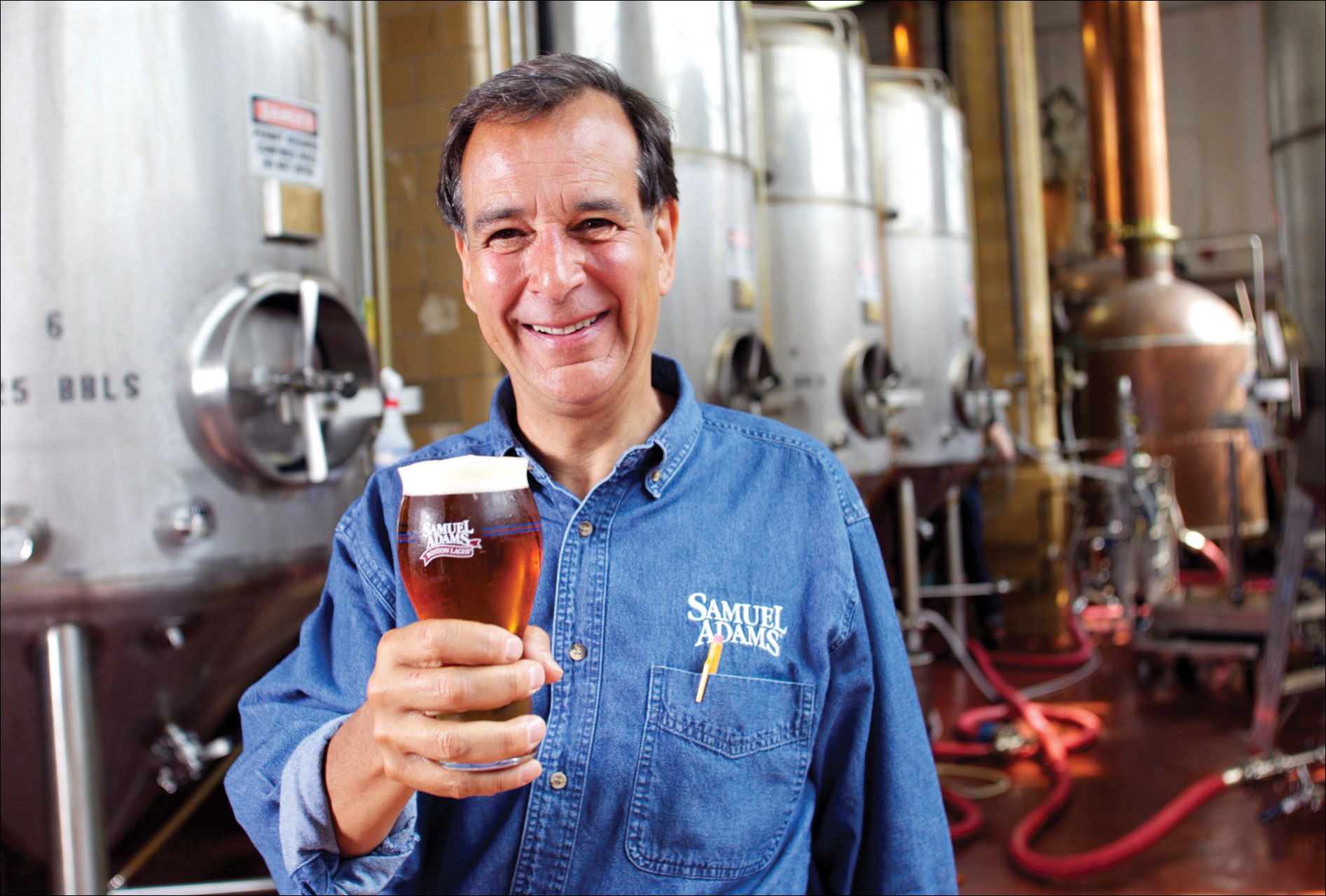 Photo shows Jim Koch holding a glass of frothing beer in a distillery.