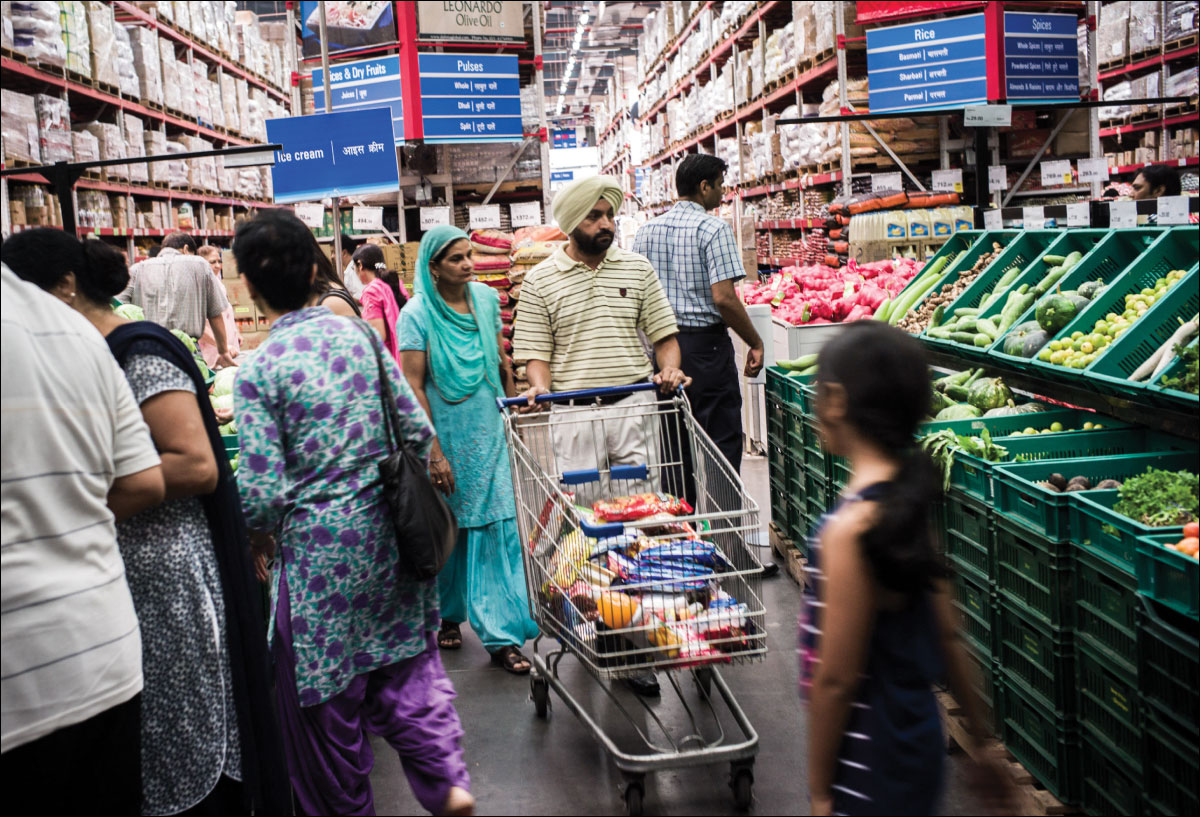 Photo shows a Walmart store in India. An Indian couple is pushing a shopping cart down one of the aisles while many other people are looking into the shelves.