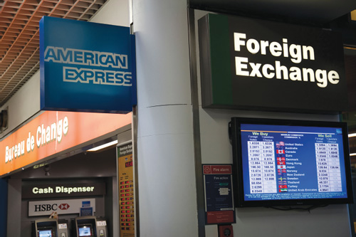 An image illustrates a signage in the Mexico City airport, which quotes exchange rates for buying and selling U.S. dollars, euros, pounds, and Canadian dollars relative to the Mexican peso. Cash dispensers of HSBC bank and American Express are visible.