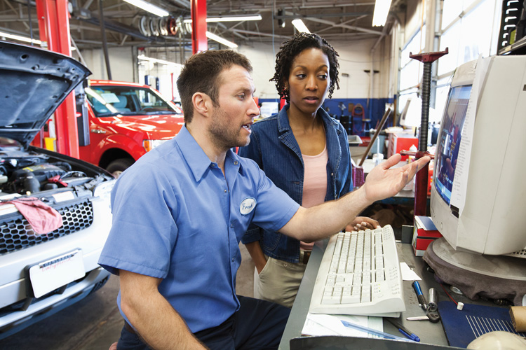 An automotive technician shows a woman a diagnostic display on a computer screen.