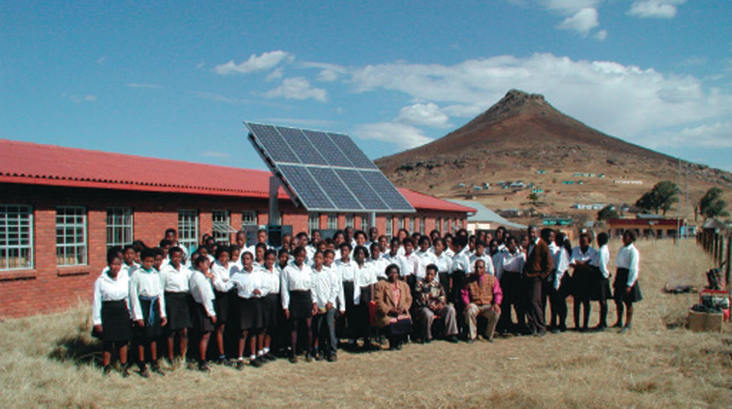Bunch of students wearing uniforms standing together with the teachers from South Africa, with a PV solar panel and a building in the background.